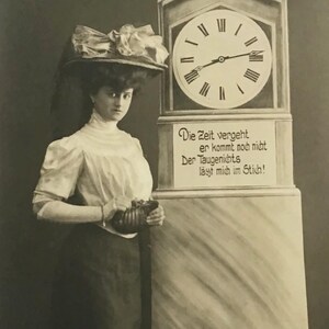 May include: Vintage black and white postcard featuring a woman in a hat standing next to a tall clock. The clock face has Roman numerals. Text on the clock reads, "Die Zeit vergeht, er kommt nob nitt, Der Taugenichts, layt mich in Sticht!"