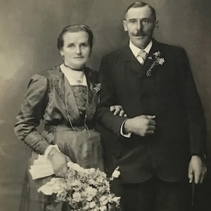 May include: Vintage black and white photograph of a formally dressed couple. The woman wears a long dress and holds a bouquet. The man is in a suit with a boutonniere. The photograph is on a slightly aged paper.