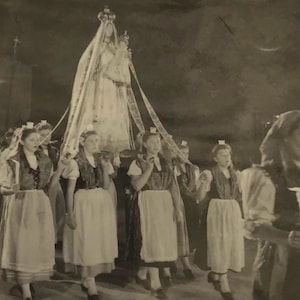 May include: Monochrome photograph depicting a religious procession. A statue of a crowned woman is carried by a group of people. The people are dressed in traditional attire, and the scene takes place indoors.
