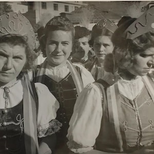 May include: A monochrome photograph showing a group of women in traditional folk dress. They are wearing elaborate headwear, vests, and blouses with puffed sleeves. The photo is mounted on patterned paper.