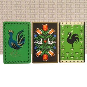 Op de afbeelding: Drie vintage speelkaarten met haan- en bloemontwerpen. De kaarten hebben een groen, zwart en crèmekleurig kleurenschema met gedetailleerde illustraties van hanen, bloemen en vogels. De kaarten hebben een decoratieve rand.