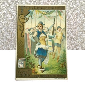 May include: Vintage advertisement card featuring a costumed figure in a blue and yellow outfit, holding a rifle. The card has text in German and Italian, with the Liebig Company logo and a decorative border. The background shows a crowd of people.