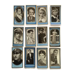 Op de afbeelding: Een verzameling vintage filmsterren verzamelkaarten. Elke kaart toont een zwart-wit portret van een acteur of actrice, tegen een blauwe achtergrond met gouden rand. Namen staan onderaan elke kaart.