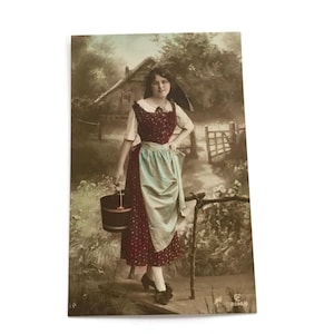 Postal antigua de una niña holandesa, coloreada a mano, con una lechera con delantal en una escena campestre.