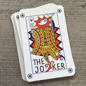May include: A pack of playing cards with a Joker card on top. The Joker card shows a jester illustration in red, yellow, and blue, holding a heart. The text "THE JOKER" is printed in black and red. The cards have a white background with blue star corner designs.