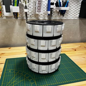 Pode incluir: Uma pilha de seis bobinas de filamento branco para impressão 3D. As bobinas são etiquetadas com o tipo de filamento e o tamanho. As bobinas estão em um tapete de corte verde.