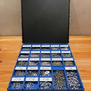 Può includere: Organizzatore in plastica nera e blu aperto, pieno di vari piccoli pezzi di ferramenta metallica. L'organizzatore ha più scomparti etichettati con viti, bulloni e rondelle. Le etichette includono dimensioni come M2, M3, M4 e M8.
