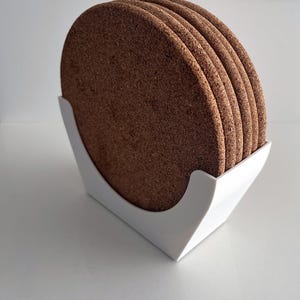 Può includere: Un set di quattro sottobicchieri circolari in sughero marrone disposti in una pila verticale. I sottobicchieri sono tenuti in posizione verticale da un supporto in plastica bianca con un bordo anteriore curvo che sostiene la forma rotonda.
