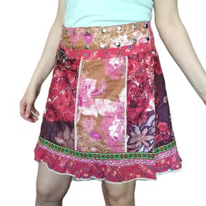 Peut inclure: Un débardeur vert menthe clair associé à une jupe en patchwork multicolore présentant divers motifs floraux dans des tons de rose, marron et violet. La jupe possède une large ceinture avec des clous en métal argenté et un ourlet à volants.