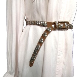 May include: A brown leather belt with silver-colored studs and a rectangular buckle. The belt is worn over a white button-down shirt. The studs are arranged in a pattern along the belt's length.