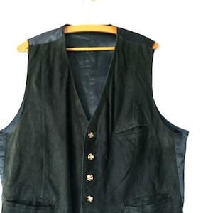 Könnte beinhalten: Dunkelgrüne Wildlederweste mit V-Ausschnitt und vier goldfarbenen Knöpfen. Die Weste hat zwei Vordertaschen und ein schwarzes Satinfutter. Sie hängt an einem Holzbügel vor weißem Hintergrund.