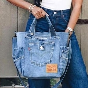 Op de afbeelding: Een blauwe denim tote tas met een voorvak met een zilveren knoop, een klein rood label en een beige patch in leerstijl. De tas heeft twee handvatten en een lange schouderriem, gemaakt van denimstof met zichtbare stiksels en afwerkingen.