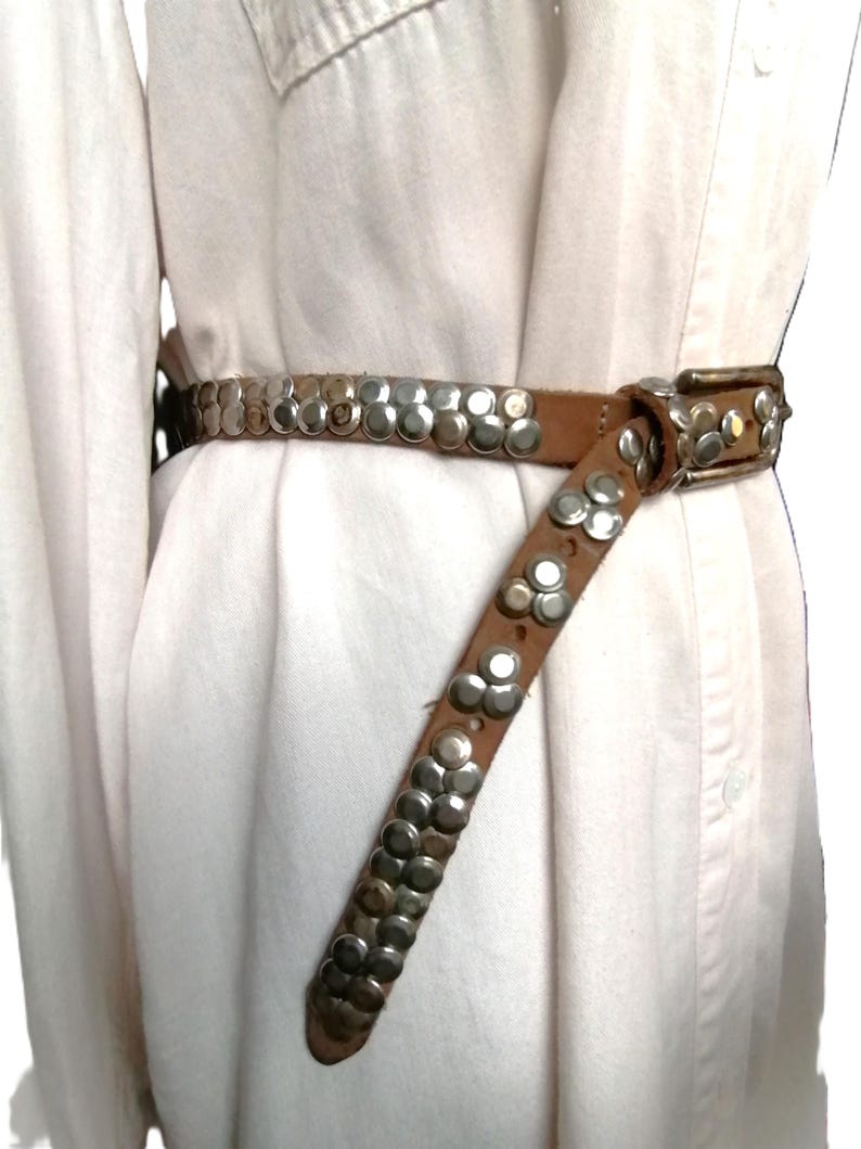 May include: A brown leather belt with silver-colored studs. The belt has a rectangular silver buckle and is worn around a white shirt. The studs are arranged in rows and clusters along the belt's length, adding a decorative element.