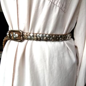 May include: A brown leather belt with silver-colored studs and a rectangular buckle. The belt is worn over a light-colored button-down shirt, creating a stylish, vintage look. The studs are evenly spaced along the belt's length.