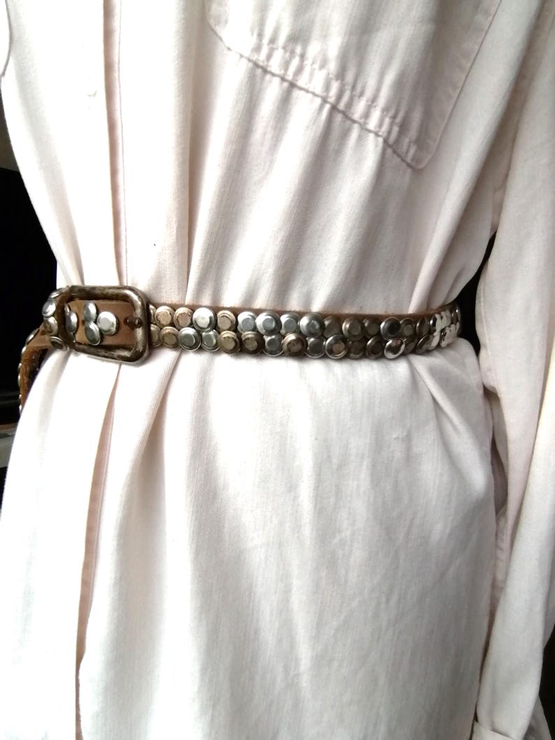 May include: A brown leather belt with silver-colored studs and a rectangular buckle. The belt is worn over a light-colored button-down shirt, creating a stylish, vintage look. The studs are evenly spaced along the belt's length.