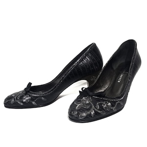 May include: A pair of black heels with a textured pattern. The shoes have a small bow on the toe and a floral design. The heels are approximately 2 inches high.