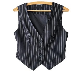 May include: A navy blue pinstripe vest with a V-neck and five dark buttons. The vest is hanging on a wooden hanger against a white background. The vertical white stripes create a classic, tailored look.