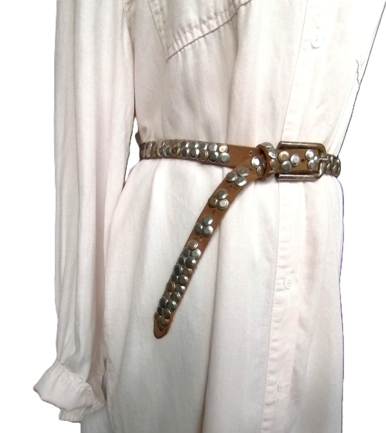 May include: A brown leather belt with silver-colored studs and a rectangular buckle. The belt is worn over a white button-down shirt. The studs are arranged in a pattern along the belt's length.