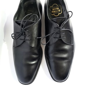 May include: A pair of black leather dress shoes with laces, displayed on a white background. The shoes have a classic design with a rounded toe and a low heel. The interior of one shoe features a gold label with the text "Centro Moda Anna Musso".
