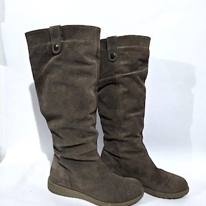May include: A pair of brown suede knee-high boots. The boots have a rounded toe, a low heel, and a button closure at the top. The suede material has a slightly worn appearance. The boots are set against a white background.