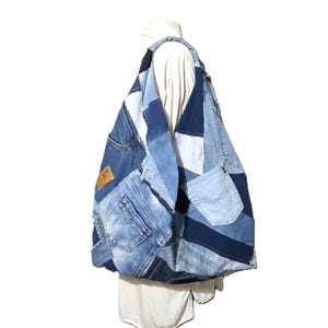 May include: A patchwork denim tote bag in various shades of blue. The bag is constructed from repurposed denim, featuring pockets and seams from jeans. A brown leather tag is visible. The bag is worn over a white shirt.