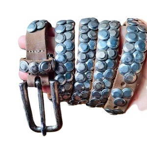 May include: A brown leather belt with silver-colored studs. The belt has a silver buckle and is studded with multiple rows of silver rivets. The belt is shown coiled, with the buckle end visible.