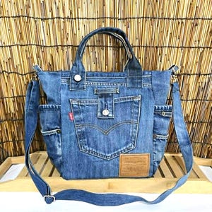 Può includere: Una borsa tote in denim blu realizzata con jeans riciclati, dotata di una tasca centrale con patta a bottone, due tasche laterali, due manici superiori e una tracolla rimovibile. Una toppa in pelle beige è cucita sul lato anteriore inferiore.