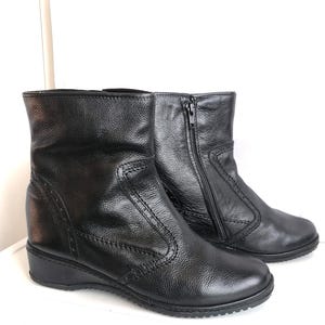 Puede incluir: Un par de botines de cuero negro con tacón de cuña bajo. Las botas tienen cierre de cremallera lateral y detalles de costuras decorativas. El cuero tiene una apariencia texturizada. Aproximadamente 15 cm de altura.