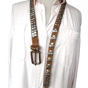 May include: A brown leather belt with silver-colored studs. The belt has a rectangular silver buckle and is displayed against a white shirt. The studs are arranged in rows and clusters, adding a decorative element to the accessory.