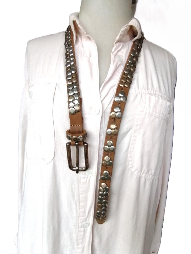 May include: A brown leather belt with silver-colored studs. The belt has a rectangular silver buckle and is displayed against a white shirt. The studs are arranged in rows and clusters, adding a decorative element to the accessory.