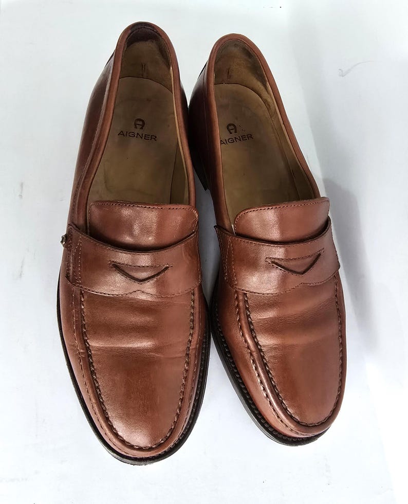 Pu&ograve; includere: Un paio di mocassini in pelle marrone con un bordo cucito e il logo Aigner sulla soletta.