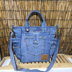 Op de afbeelding: Een blauwe denim tas gemaakt van gerecyclede jeans, voorzien van twee handvatten en een afneembare schouderriem. De voorzijde heeft een centraal vak met een knoopsluiting, een klein rood Levi's label en een bruin lederlook merklabel.