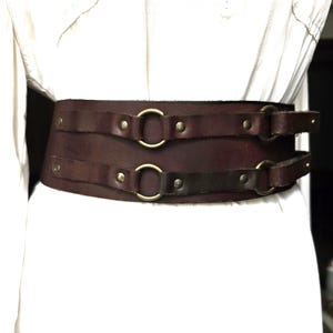 May include: A wide, dark brown leather belt with two horizontal straps and brass-colored metal rings and rivets. The belt is designed to be worn around the waist. The leather appears to be distressed, giving it a vintage look.