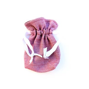 May include: A small, drawstring bag in a diamond pattern, primarily in shades of burgundy and white. The bag is gathered at the top with white drawstrings tied in a bow. The bag is likely used for small gifts or storage.