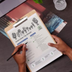 May include: A wooden clipboard displays an "Interview Tip Sheet" with microphone illustrations. A hand holds a pen, pointing to the sheet. The scene includes a laptop, a plant, and a decorative glass container, all on a dark wooden surface.