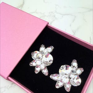 May include: A pair of sparkling, flower-shaped earrings with clear crystals, presented in a pink box with a black interior. The earrings have a central, large crystal surrounded by smaller ones.