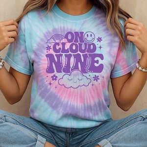May include: Tie-dye t-shirt in shades of blue, pink, and purple. The shirt features the phrase "ON CLOUD NINE" in purple, along with a smiley face, rainbow, cloud, and floral graphics. The model is wearing blue jeans.