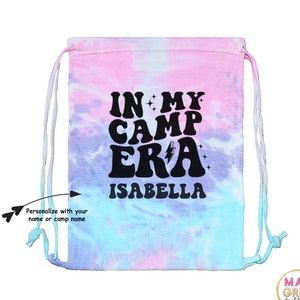 May include: A pink, blue, and white tie-dye drawstring bag with the text "IN MY CAMP ERA" and the name "ISABELLA" in black. The bag is personalized with a name or camp name.