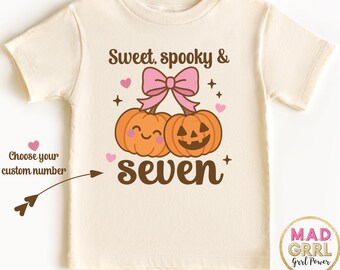 May include: Cream-colored t-shirt featuring a Halloween-themed graphic. The design includes two smiling pumpkins, a pink bow, hearts, and the text "Sweet, spooky & seven." The shirt has short sleeves and a round neckline.