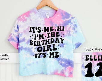 May include: A tie-dye crop top with the text "It's Me, Hi I'm The Birthday Girl It's Me" in black. The back of the shirt has the name "Ellie" and the number "12" in black.