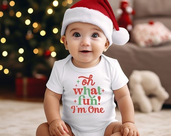 Puede incluir: Un bebé con un gorro de Papá Noel rojo y blanco y un body blanco con el texto "Oh what fun! I'm One" en rojo y verde. El bebé está sentado sobre una superficie clara con un árbol de Navidad borroso en el fondo.