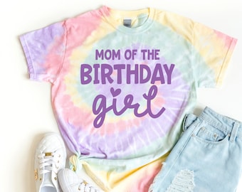 May include: A tie-dye t-shirt with the text "Mom of the Birthday girl" in purple. The shirt is on a white background with a pair of white sneakers and a pair of light blue denim shorts.
