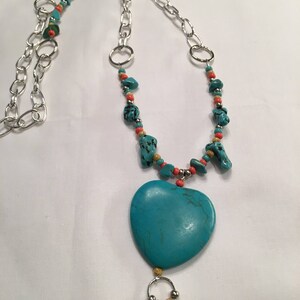 May include: A turquoise and silver necklace with a large turquoise heart pendant. The necklace is made with turquoise stones, silver chain, and small orange and gold beads.