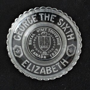 May include: A clear glass plate with a scalloped edge. The plate features a raised design of the text "George the Sixth Elizabeth" and a royal crest with the text "Royal Visit Souvenir Canada 1939".