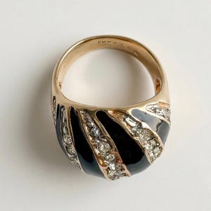 May include: Gold-toned ring with a black enamel band and clear crystal accents. The ring features a wide, oval design with a series of black and crystal stripes. The band is smooth and polished, creating a striking contrast with the textured details.