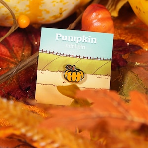 May include: A mini pin depicting a cartoon pumpkin with a brown stem, set against a green field and a blue sky. The pin is on a card that reads "Pumpkin mini pin". The image is surrounded by autumn leaves and gourds.