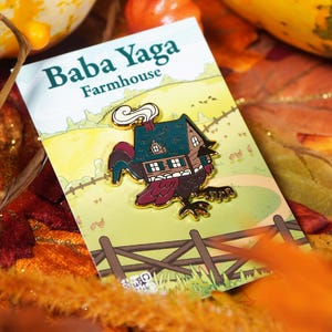 Pode incluir: Um alfinete de esmalte Baba Yaga Farmhouse. O alfinete apresenta uma casa com corpo de galo, telhado verde e chaminé fumegante. O alfinete está em um cartão com o texto "Baba Yaga Farmhouse" e uma paisagem de desenho animado.