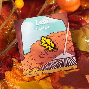 May include: A mini pin with a yellow leaf design and black outlines, set against a fall-themed illustration of a leaf pile and rake. The pin is on a card that reads "Leaf mini pin". The background includes fall foliage.