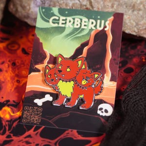 Puede incluir: Un pin de esmalte rojo que representa a Cerbero, el perro de tres cabezas, con detalles amarillos y un collar de púas. El pin está sobre una tarjeta con el nombre "CERBERUS" y un fondo de un paisaje en llamas. También se ven un hueso y una calavera.