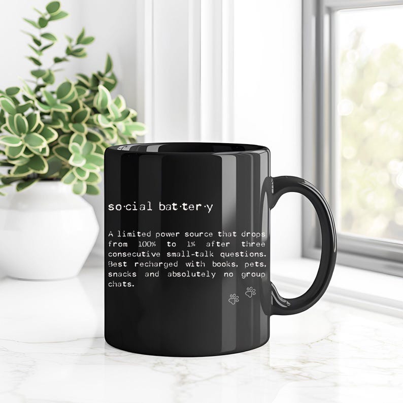 May include: Black ceramic mug with the text "social battery" and a humorous description about social interaction. The mug also features paw prints. A perfect gift for introverts.
